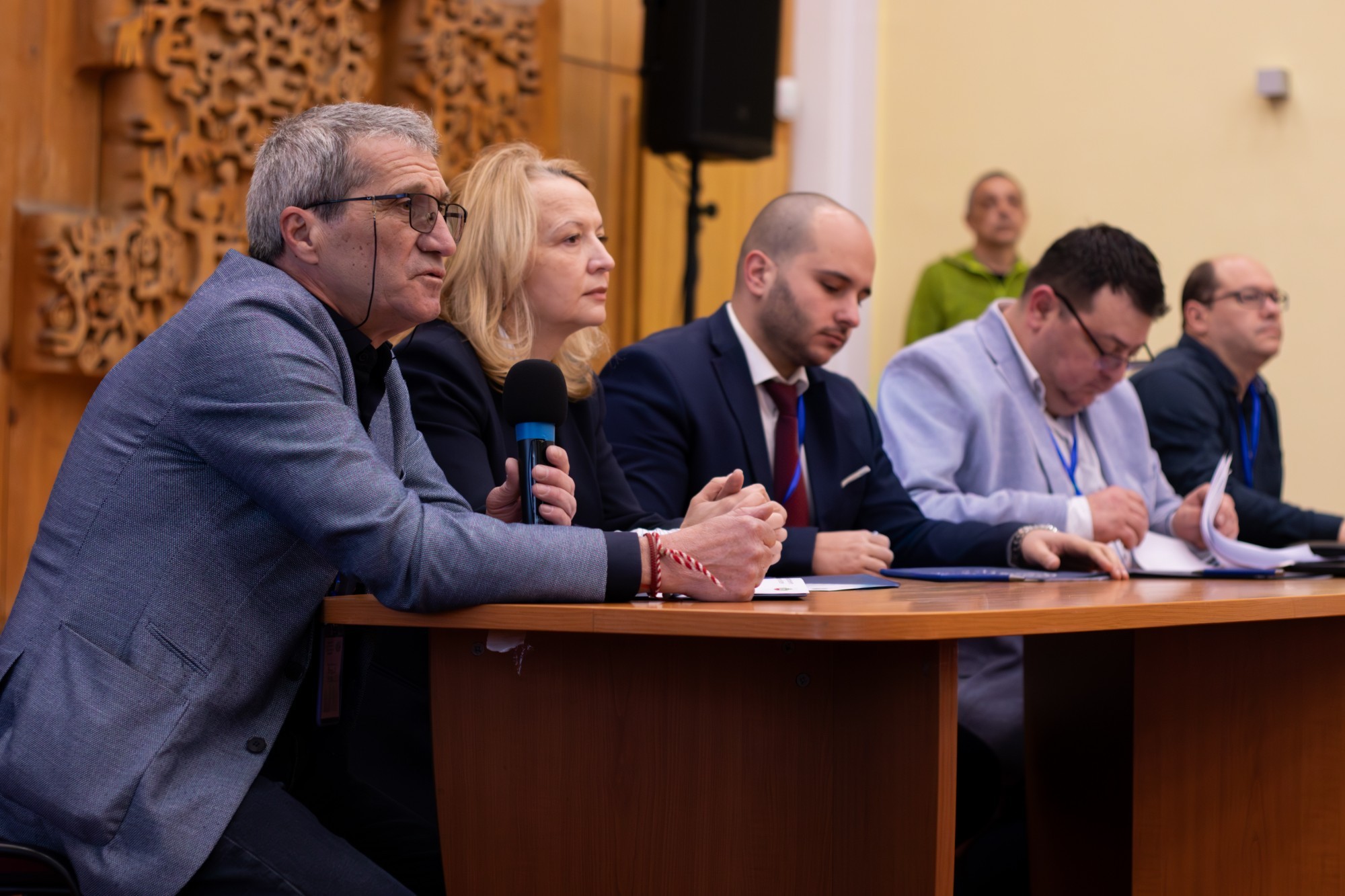 Президиум на среща на професионални домоуправители: мъж с микрофон говори, до него седят участници в залата